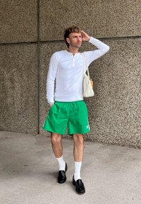 Weißes Langarmhemd, grüne Shorts, schwarze Slipper und weiße Socken. Bemerkenswerte Details sind ein kleines Markenlogo und eine weiße Handtasche.