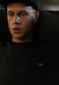 Maglietta atletica nera con motivo a rilievo e piccolo logo "ICIW" sul petto, indossata da una persona con capelli rasati, seduta contro uno sfondo scuro.