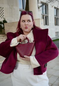 Bordó kézitáska strukturált, háromszög alakú, rövid fogantyúval, egy krémszínű pulóvert és bordó kabátot viselő személy által tartva.