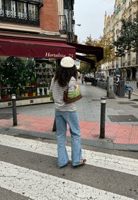 Prugasta majica dugih rukava u zelenoj i žutoj boji, plave široke traperice, bež beretka i smešak torba, stojeći na pešačkom prelazu.
