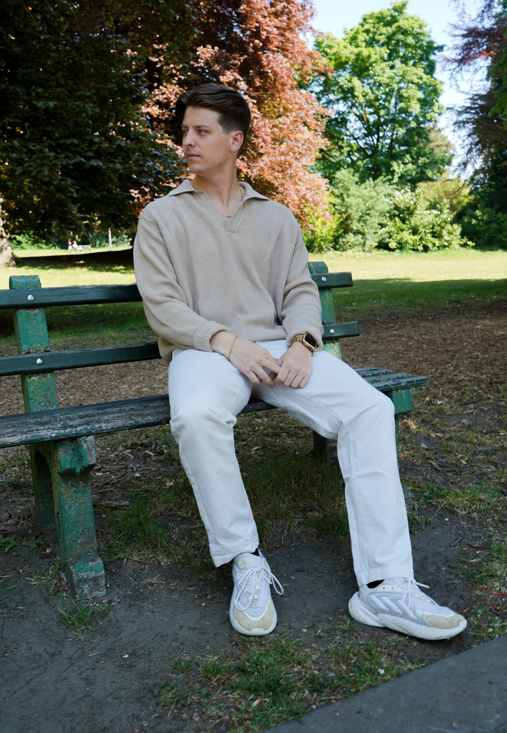 Pull en maille beige clair avec un col, pantalon blanc et baskets gris-blanc. Personne assise sur un banc de parc vert entouré de verdure.