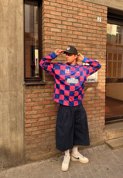 Joven con sudadera de Nike a cuadros azul y rosa, gorra negra, pantalones cortos azul marino y zapatillas blancas, de pie junto a una pared de ladrillos al aire libre.