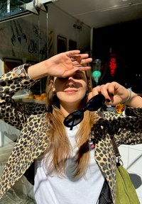 Casaco de pele de leopardo, t-shirt branca, mãos a proteger os olhos da luz solar, segurando óculos de sol pretos. Iluminação brilhante com tons quentes.