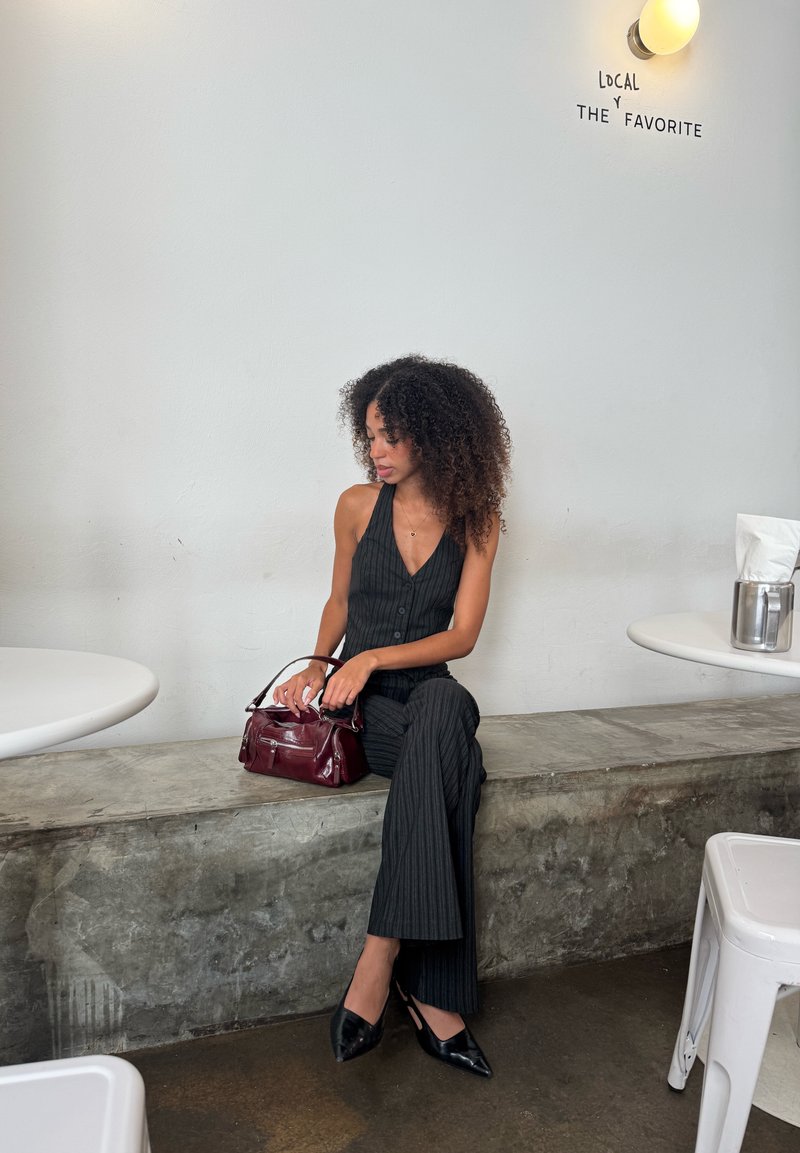 Macacão preto riscado com decote em V, combinado com sapatos pretos de bico fino e uma bolsa bordeaux. Parede lisa ao fundo.
