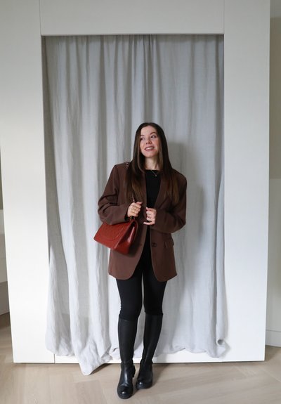 Blazer marrón sobre una blusa negra, combinado con leggings negros y botas altas negras. Sosteniendo un bolso acolchado rojo. Fondo de cortina neutral.
