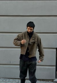 Jachetă maro scurtă cu finisaj texturat și nasturi, purtată peste un top bleumarin. Asortată cu pantaloni largi de culoare închisă, pe un fundal gri.