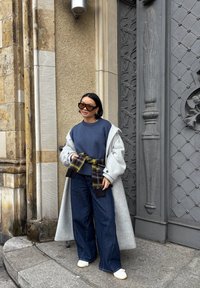 Szary, oversize płaszcz na granatowym swetrze, zapinany paskiem w kratę. Połączony z szerokimi, ciemnymi dżinsami i białymi trampkami. Brązowe okulary przeciwsłoneczne.