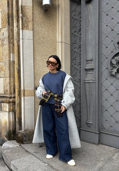 Abrigo gris de gran tamaño sobre un suéter azul marino, ceñido con un cinturón de patrón a cuadros. Combinado con vaqueros oscuros de pierna ancha y zapatillas blancas. Gafas de sol marrones.