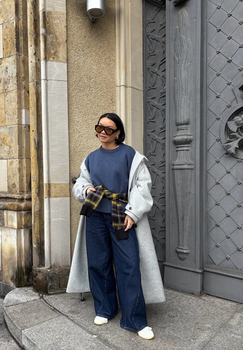 Palton gri supradimensionat peste un pulover bleumarin, prins cu o curea în model în carouri. Asortat cu blugi evazați închiși la culoare și adidași albi. Ochelari de soare maro.