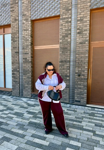 Chándal burdeos con rayas blancas, pantalones de corte holgado y una camisa blanca abotonada. Bolso negro, gafas de sol y fondo de ladrillo.