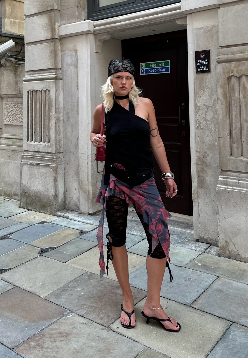 Black sleeveless top, floral patterned wrap skirt in shades of grey and red, black lace leggings, and black heeled sandals. Red handbag and jewellery.