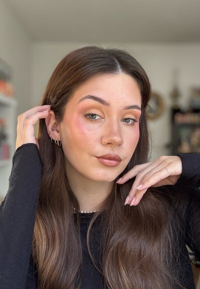 Joven mujer con largo cabello castaño, sutiles pecas, anillo de septum dorado y pendientes, vestida con camiseta negra, tocándose el cabello y la barbilla en un ambiente interior.