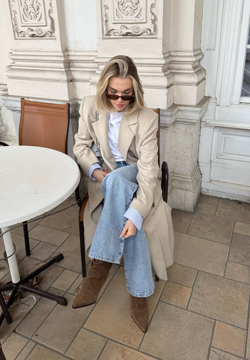 Femme aux cheveux blonds portant un manteau beige, une chemise blanche, un jean bleu, des bottes marron et des lunettes de soleil, assise à une table de café en plein air avec un mur en pierre orné en arrière-plan.