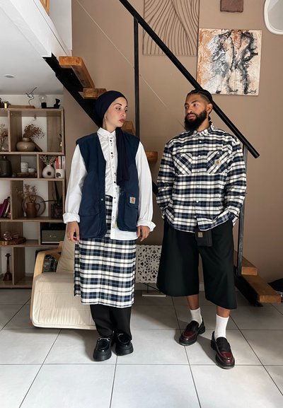 Dos personas están de pie una al lado de la otra. La mujer lleva un chaleco azul marino sobre una camisa blanca y una falda de cuadros. El hombre lleva una camisa de cuadros, pantalones cortos y zapatos oscuros.