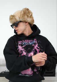 Μαύρη oversized φούτερ μπλούζα με γραφικό τύπωμα που απεικονίζει αστραπές, φορεμένη με καπέλο από συνθετική γούνα σε μπεζ απόχρωση και σκούρα γυαλιά ηλίου. Απλό, μοντέρνο στυλ.