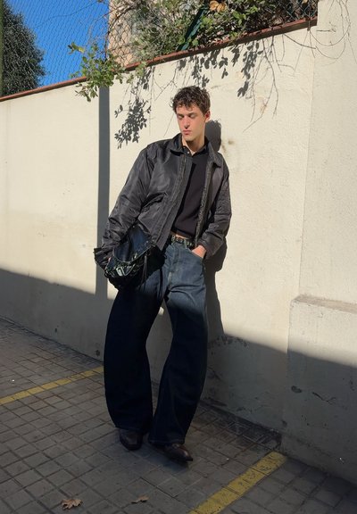 Chaqueta bomber negra sobre un polo negro, pantalones de mezclilla azul holgados y zapatos marrones. Sosteniendo una bolsa brillante de color verde oscuro. Entorno al aire libre iluminado por el sol.