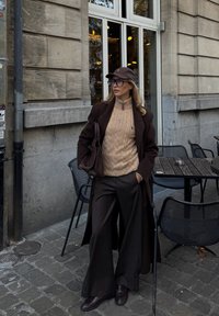 Brun uldfrakke, beige kabelstrikket sweater, brede brune bukser, brun kasket, stor håndtaske, stående på brosten nær udendørs café.