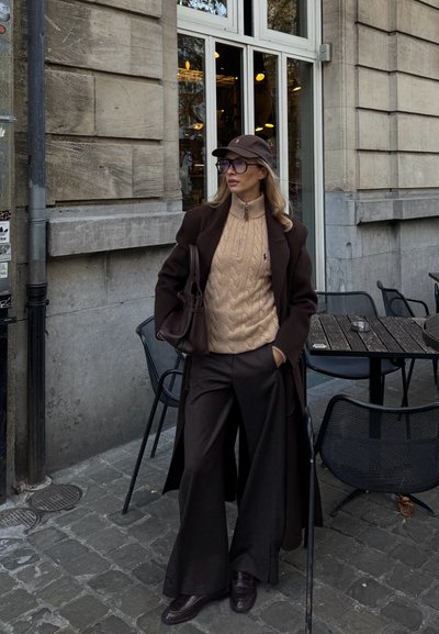 Abrigo de lana marrón, suéter beige de punto trenzado, pantalones marrones de pierna ancha, gorra marrón, bolso grande, de pie sobre adoquines cerca de una cafetería al aire libre.