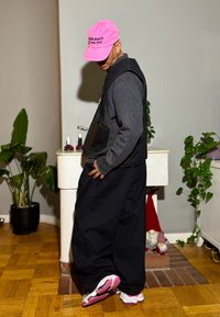 Vêtements superposés noirs et gris avec une casquette rose ornée de texte. La personne porte un pantalon noir surdimensionné et des baskets multicolores.