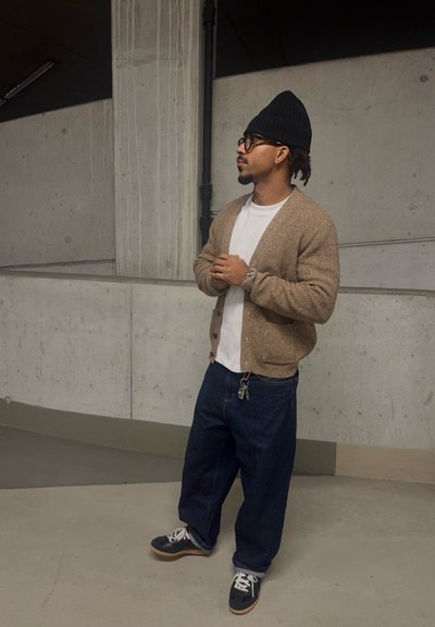 Hombre que lleva un gorro negro, gafas, cárdigan beige, camisa blanca, vaqueros anchos azules y zapatillas negras, está de pie con las manos entrelazadas en un espacio interior de concreto.
