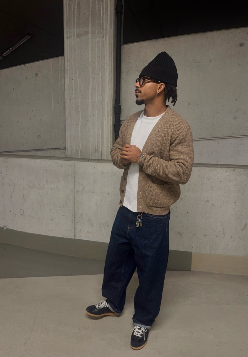Homme portant un bonnet noir, des lunettes, un cardigan beige, une chemise blanche, un jean large bleu et des baskets noires, se tenant les mains jointes dans un espace intérieur en béton.