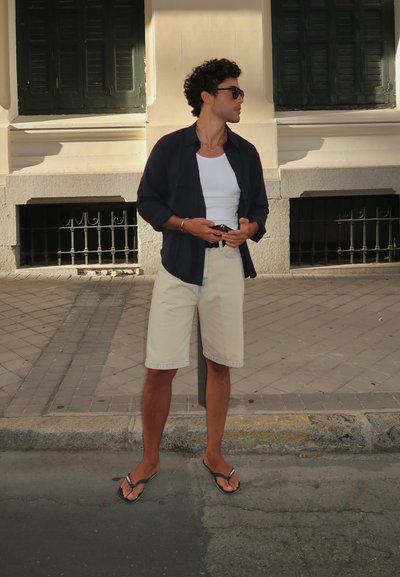 Conjunto casual que incluye una camisa abierta de color azul marino oscuro, una camiseta blanca, pantalones cortos beige y chanclas negras, de pie en una acera.