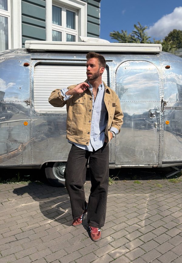 Homme en veste beige et pantalon noir debout sur le trottoir devant un camping-car argenté brillant sous un ciel bleu.