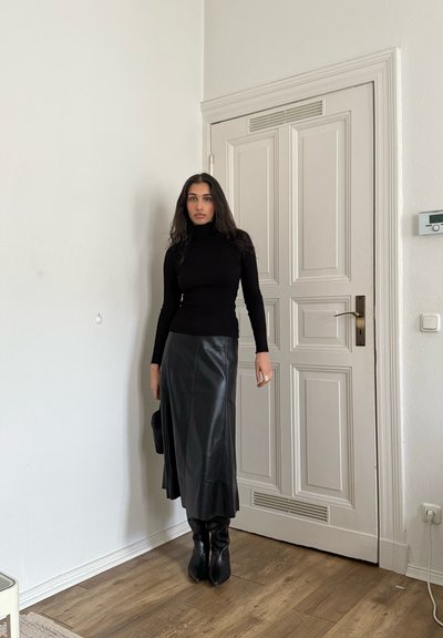 Suéter de cuello alto acanalado negro, falda larga de cuero negro de talle alto, botas negras hasta la rodilla, con fondo de una pared y puerta blancas.