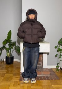 Ruskea toppatakki, jossa huppu, musta paita alla, väljät siniset farkut ja moniväriset lenkkarit. Sisätilassa vihreiden kasvien kanssa.