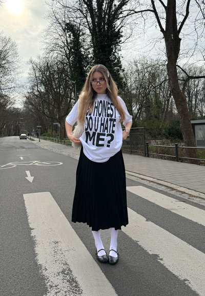 Camiseta blanca de gran tamaño con texto negro en negrita, falda midi plisada negra, calcetines blancos y zapatos negros, de pie en un paso de peatones.