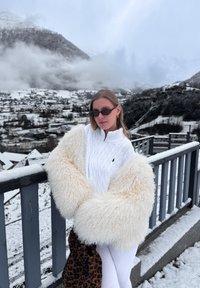 Femme portant un pull blanc et une grande veste crème et duveteuse, debout sur un balcon enneigé avec un village de montagne et des nuages en arrière-plan.