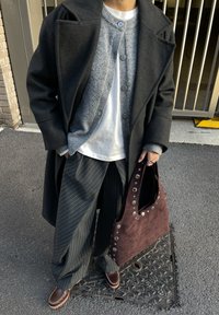 Personne portant un manteau long gris foncé, un cardigan boutonné gris, une chemise blanche, un pantalon large à fines rayures, des chaussures marron, tenant un sac à main marron clouté sur le trottoir.