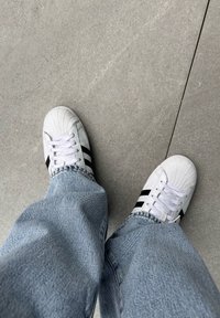 Adidași albi cu dungi negre, vârf texturat și șireturi albe. Purtați cu blugi largi de denim albastru deschis pe o suprafață gri.