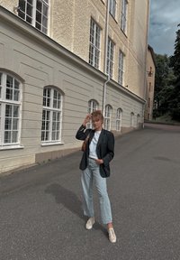 Μαύρο σακάκι πάνω από ένα ανοιχτό μπλε πουκάμισο, ανοιχτό μπλε ψηλόμεσες τζιν παντελόνες και μπεζ αθλητικά παπούτσια. Καφέ τσάντα ώμου. Ο φόντος περιλαμβάνει ένα χαμηλό κτίριο με μεγάλες πόρτες.