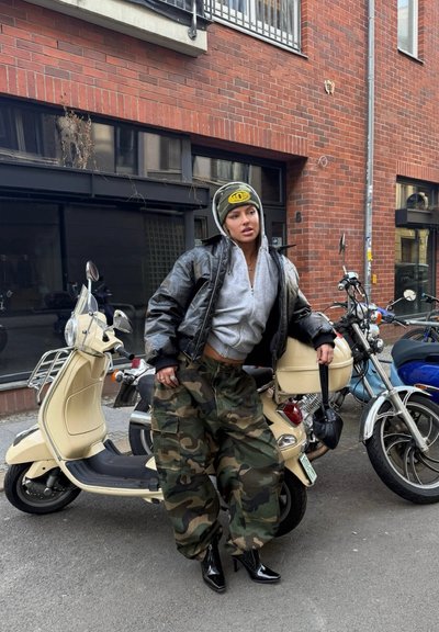 Chaqueta bomber de piel negra, sudadera gris con capucha, pantalones cargo de camuflaje y tacones altos negros. Scooter beige y motocicletas aparcadas de fondo.