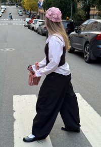 Naine, kellel on peas roosa kudumismüts, valge pluus ja mustad laiade püksteh, hoiab roosat kotti ja telefoni, kui ületab linnatänavat ülekäigurajal.