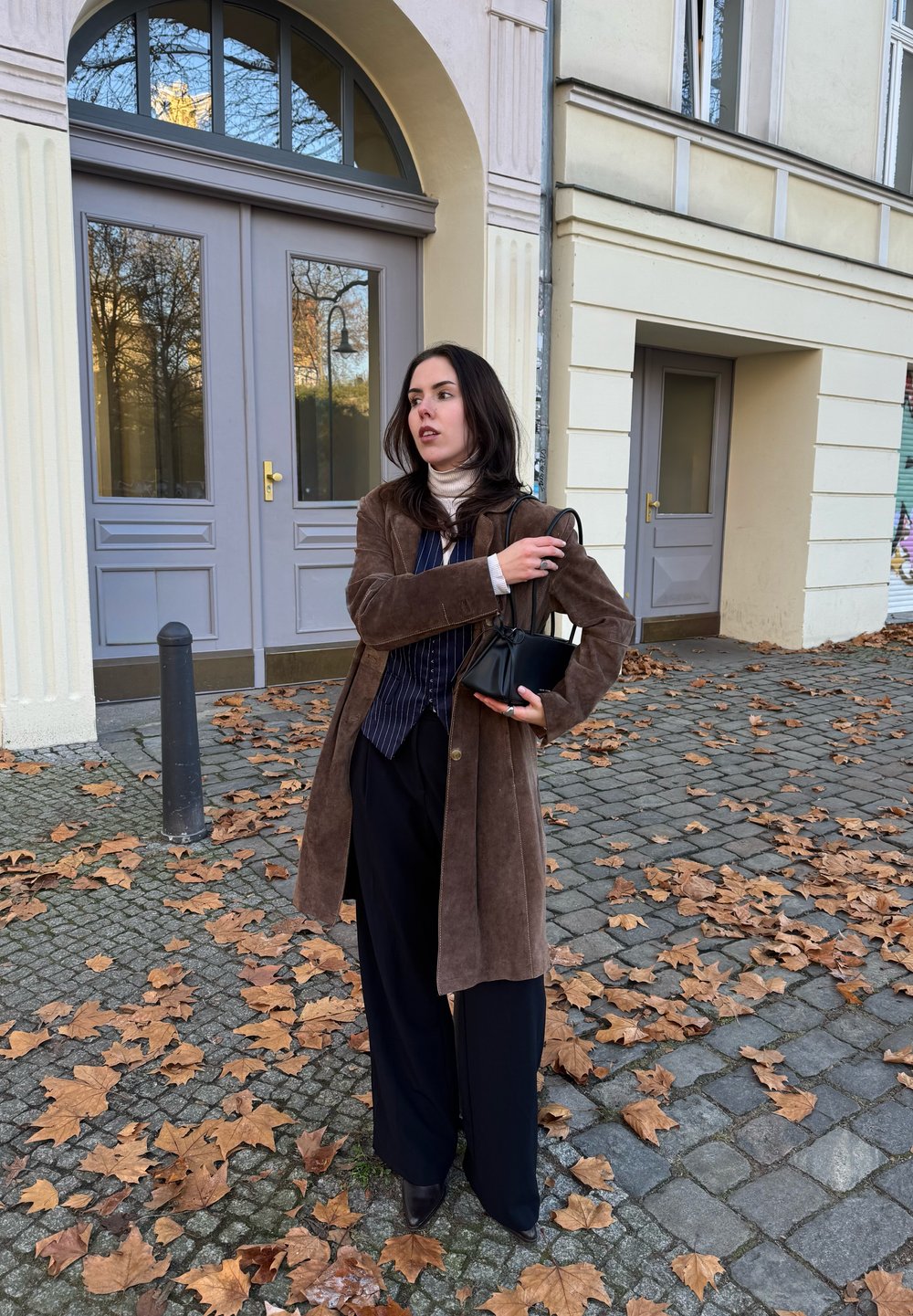 Brūna zamšādas mētelis, melna svītraina krekls, melnas plataji bikses, turot mazu melnu somiņu. Bruģa segums ar rudens lapām.