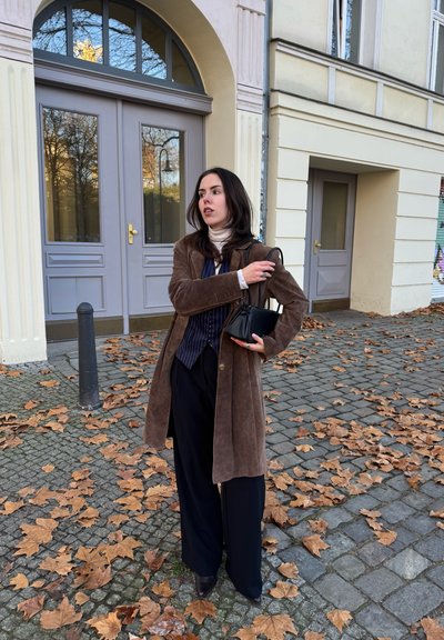 Abrigo de ante marrón, camisa negra con rayas finas, pantalones anchos negros, sosteniendo un bolso pequeño negro. Suelo de adoquines con hojas de otoño.
