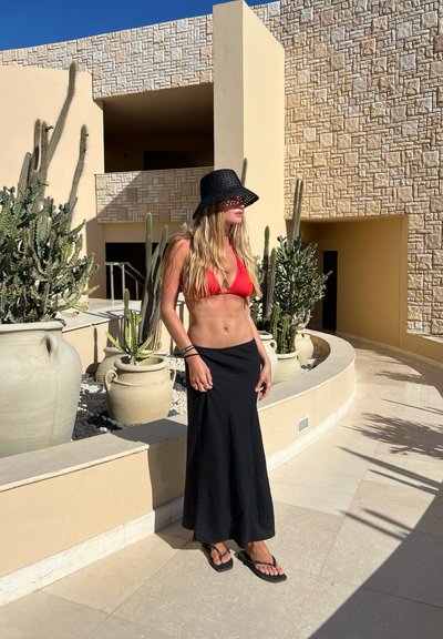 Top de bikini rojo, falda larga negra y sombrero de paja negro; de pie en un camino de piedra con cactus y macetas decorativas al fondo.