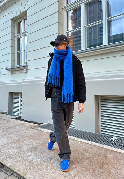 Chaqueta de plumas negra, cuello de tortuga negro, bufanda de lana azul, jeans grises y zapatillas azules con suela de goma, caminando por la acera cerca de un edificio de color claro.