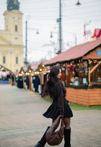 Sac à bandoulière marron surdimensionné, jupe plissée noire et bottes noires montant jusqu'aux genoux. En arrière-plan, des étals en bois et des lumières décoratives.