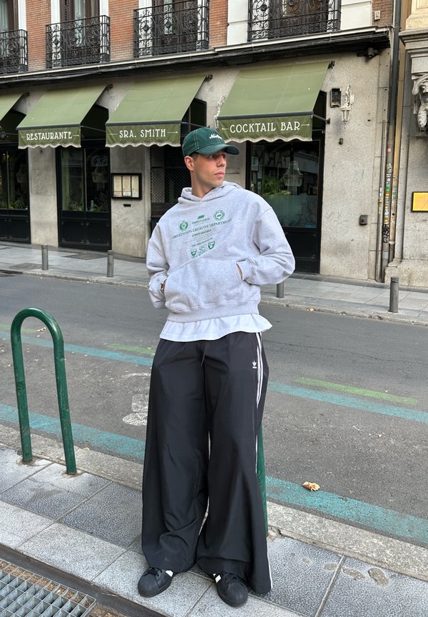 Gray hoodie with green graphic, layered over a white shirt. Loose black track pants with white stripes, and black sneakers. Urban setting.