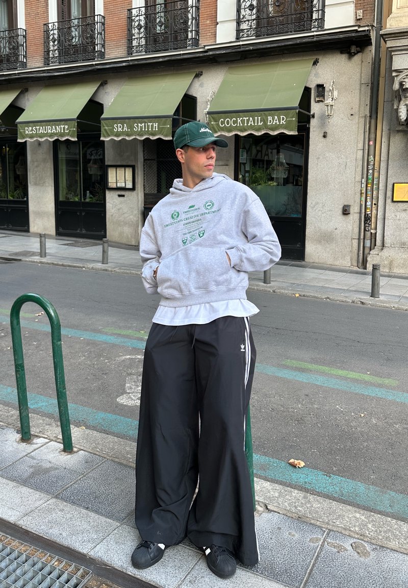 Sweat à capuche gris avec motif vert, porté sur un t-shirt blanc. Pantalon de jogging noir ample avec des bandes blanches, et baskets noires. Cadre urbain.