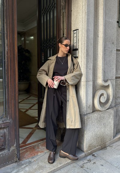 Larga gabardina beige sobre una camisa negra y pantalones negros de pierna ancha, sosteniendo una taza blanca. Zapatos marrones tipo slip-on y joyería minimalista de plata.