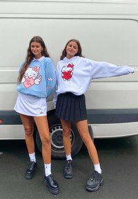Dwie kobiety noszą pastelowe bluzy z grafiką Hello Kitty, jedna w jasnoniebieskim kolorze, a druga w białym, zestawione z czarnymi spódnicami i masywnymi butami.