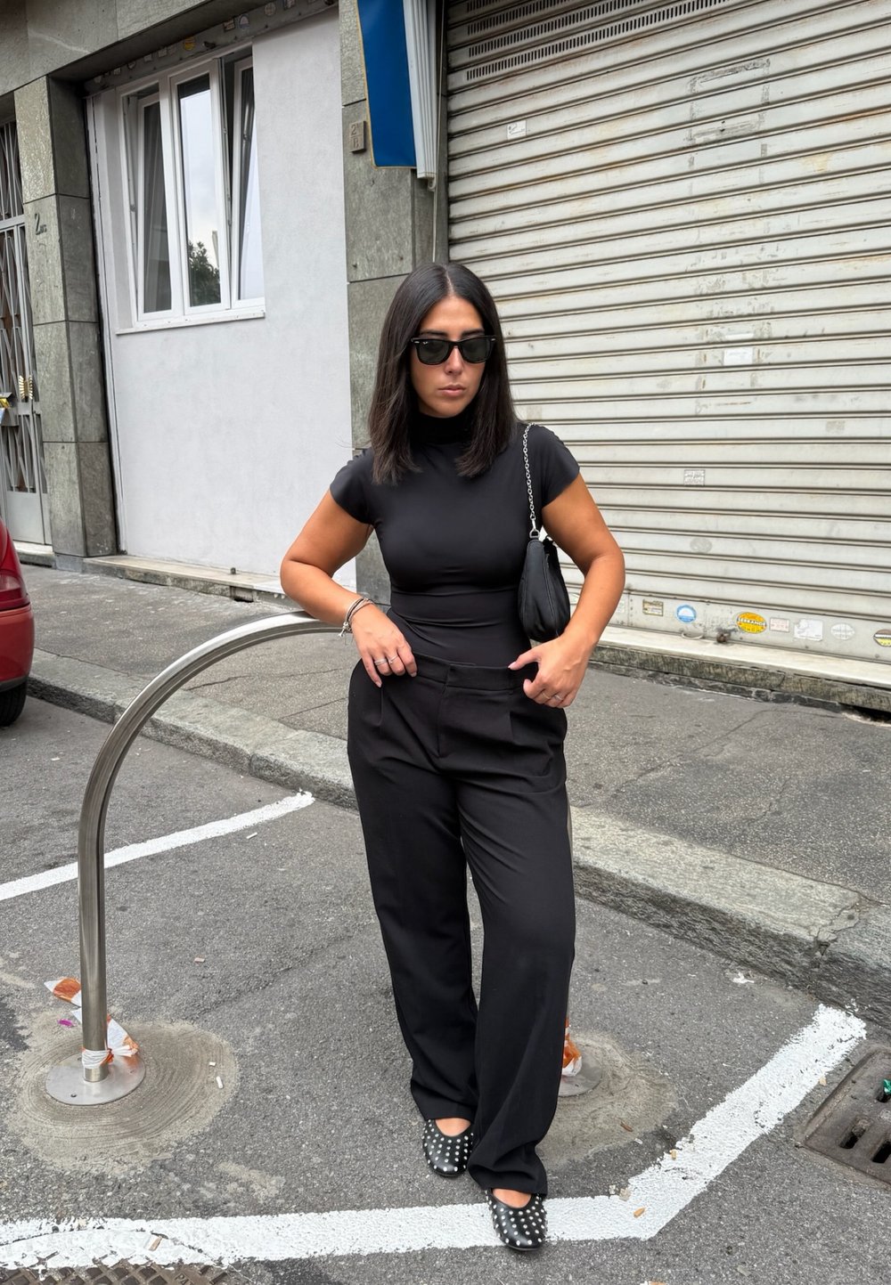 Donna in abbigliamento nero e occhiali da sole, in piedi con le mani nelle tasche su una strada urbana vicino a un portabici in metallo e la serranda di un negozio chiuso.