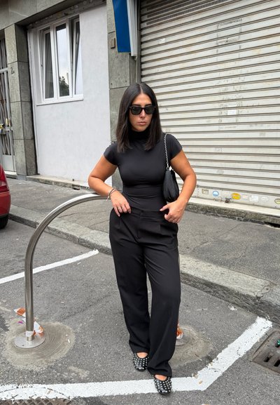 Mujer con atuendo negro y gafas de sol de pie con las manos en los bolsillos en una calle urbana, cerca de un soporte metálico para bicicletas y la persiana cerrada de una tienda.
