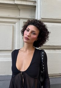 Abito nero trasparente con scollatura a V profonda e dettaglio con fiocco sul davanti. Capelli ricci, accessori minimi e una borsa nera con texture e borchie.