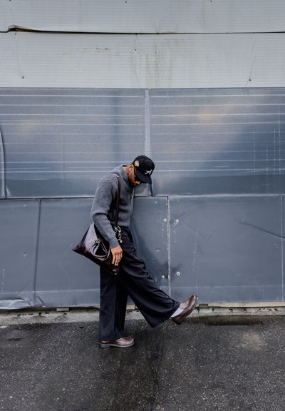 Hombre con suéter gris y gorra negra estira la pierna mientras está de pie sobre un pavimento mojado frente a una pared pintada de gris.
