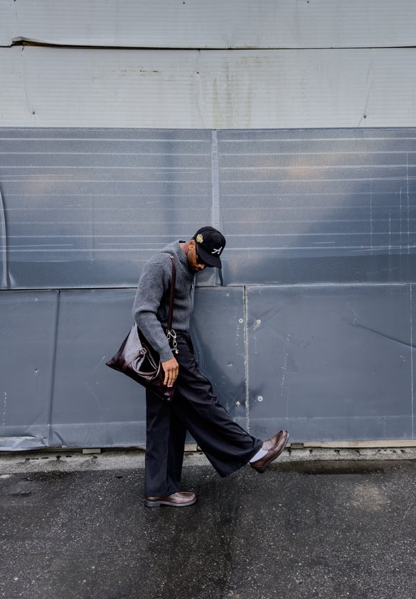 Homme portant un pull gris et une casquette noire étire la jambe en se tenant sur un trottoir mouillé devant un mur peint en gris.
